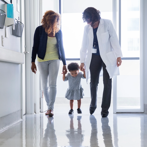 Mother and pediatrician holding hands of toddler while walking in hospital corridor.