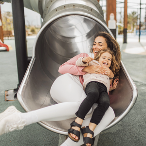 Young happy mother with her daughter having fun in tube slide on playground.