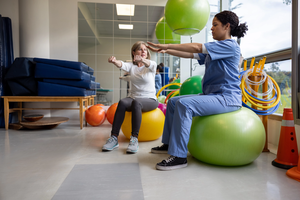 Physical therapist showing a Latin American woman a stretching exercise for her recovery at a rehab center while sitting on a fitness ball