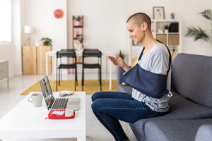 Woman in an arm sling using a laptop while having a video call with her doctor sitting on a sofa at home.