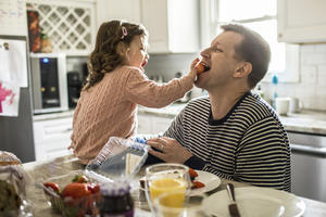 Toddler girl feeding her father a strawberry in kitchen